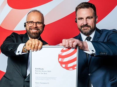Dvaja muži vo formálnom oblečení – vľavo v smokingu s okuliarmi, vpravo v tmavomodrom obleku – spoločne držia v oboch rukách zarámovaný certifikát o udelení ocenenia. Na certifikáte je výslovne uvedené „Víťaz Red Dot 2025 Best of the Best“ udelený zariadeniu „Vasco Translator V1“. V pozadí je viditeľné rozsiahle, dynamické grafické pozadie s abstraktnými červenými a bielymi vlnovými vzormi.