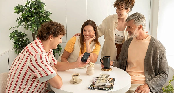 A photograph of four people gathered around a small, round white table in a bright, minimalist room. On the left, a man with curly brown hair wearing a red-and-white striped shirt holds up a dark handheld electronic device showing a numeric keypad on its screen. Seated in the middle, a woman in a yellow t-shirt and cream vest rests her chin on her hand, smiling. Behind her stands a woman with curly hair in a beige shirt, while a grey-haired man with a beard sits on the right wearing a grey cardigan and holding a black mug. The table is set with coffee cups, newspapers, and a small decorative vase under bright, soft lighting.