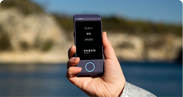 Záber zblízka na ruku, ktorá drží fialové elektronické prekladové zariadenie Vasco na pozadí jasného vonkajšieho prostredia. Na obrazovke sa zobrazujú pozdravy „Hello!“, „¡Hola!“ a logo „VASCO Translator“. V pozadí je rozmazaný pohľad na slnečnú pobrežnú krajinu s modrou vodou a svetlými skalnatými útesmi pod jasnou oblohou. Osoba držiaca zariadenie má na rukáve svetlosivú alebo striebornú textúrovanú bundu. 