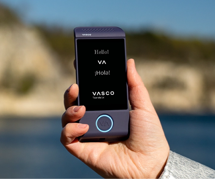 Detailný záber na ruku, ktorá drží fialový elektronický prekladač Vasco Translator M4 na jasnom exteriérovom pozadí. Na displeji sa zobrazujú pozdravy „Hello!“, „¡Hola!“ a logo „VASCO Translator“.